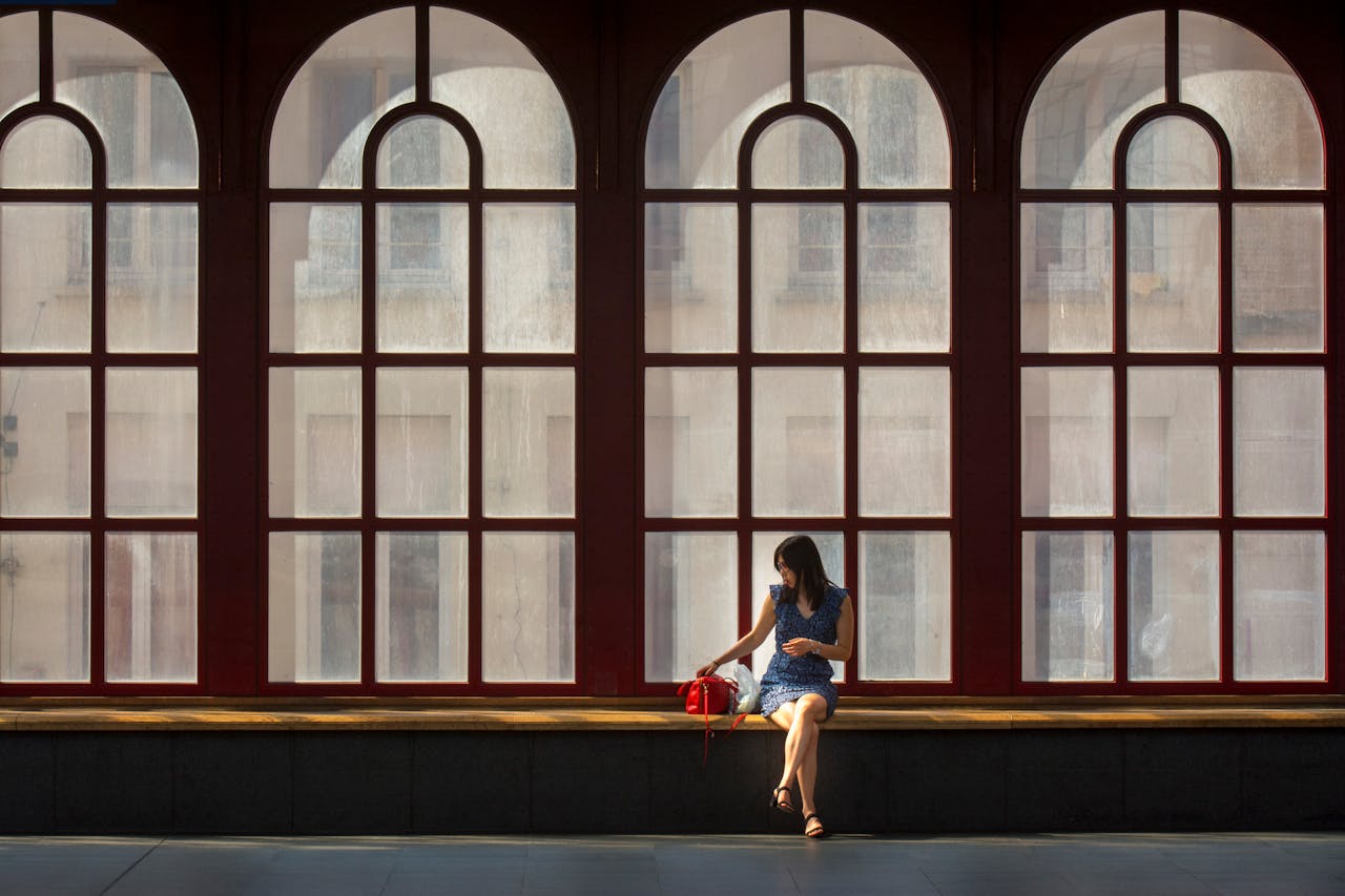 A woman sits alone in the Antwerp railway station, silhouetted by large arched windows.
