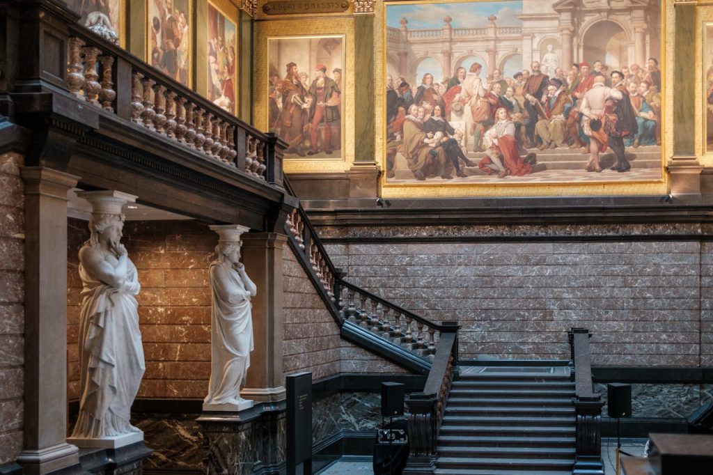 Elegant museum interior showcasing classical statues and grand staircase leading to large historical paintings.